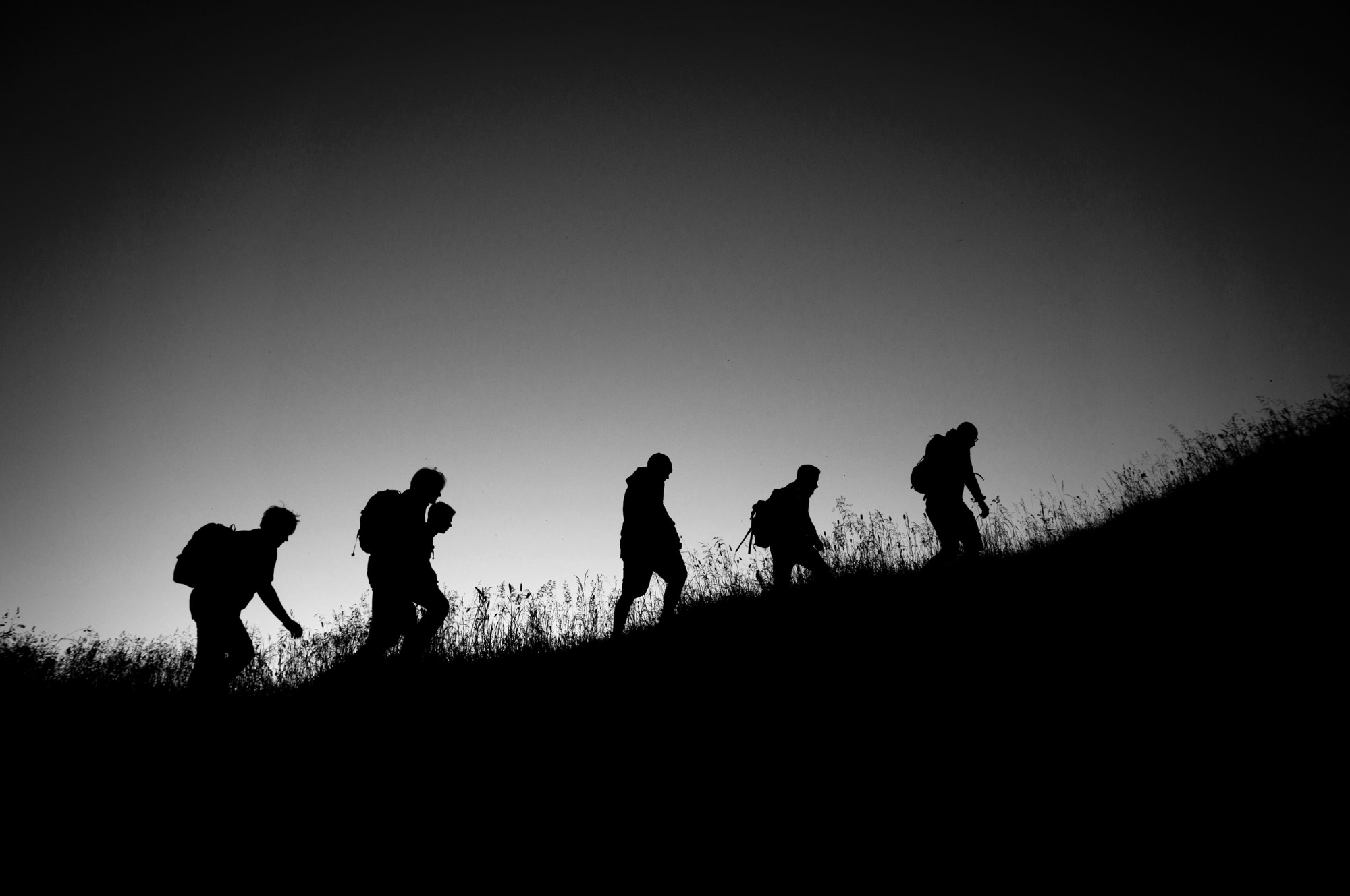 Six walkers in silhouette climbing a grassy hill at dusk
